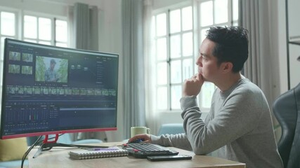 Asian Video Editor Man In Long Sleeved T-Shirt Thinking While Using Desktop Computer For Editing Video At Home.
