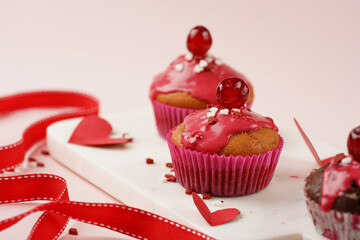 Three homemade white and chocolate muffins with red frosting and heart-shared red and white sugar decoration on marble board on pink surface