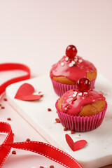 Three homemade white and chocolate muffins with red frosting and heart-shared red and white sugar decoration on marble board on pink surface