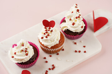 Three homemade white and chocolate muffins with white creamy frosting and heart-shared red and white sugar decoration on marble board on pink surface