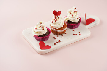 Three homemade white and chocolate muffins with white creamy frosting and heart-shared red and white sugar decoration on marble board on pink surface