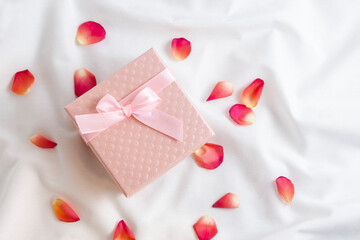 Top view of pink gift box with rose petals on white bed sheets. Romantic Valentines Day surprise and present