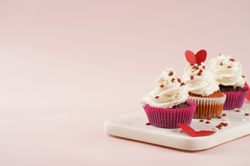 Three homemade white and chocolate muffins with white creamy frosting and heart-shared red and white sugar decoration on marble board on pink surface, copy space