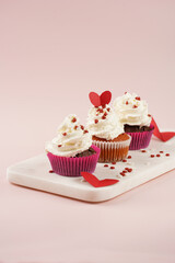 Three homemade white and chocolate muffins with white creamy frosting and heart-shared red and white sugar decoration on marble board on pink surface