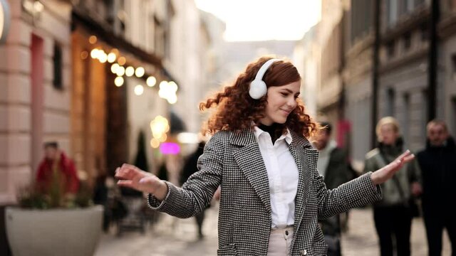 Slow motion of happy young woman in wireless headphones dancing singing outdoors in city street having fun alone. Attractive young red hair woman listening to music in headphone