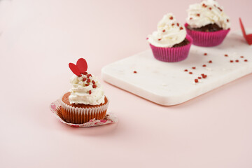 Three homemade white and chocolate muffins with white creamy frosting and heart-shared red and white sugar decoration on marble board on pink surface