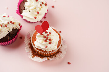 Three homemade white and chocolate muffins with white creamy frosting and heart-shared red and white sugar decoration on marble board on pink surface