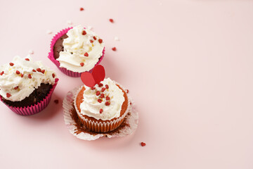 Three homemade white and chocolate muffins with white creamy frosting and heart-shared red and white sugar decoration on marble board on pink surface