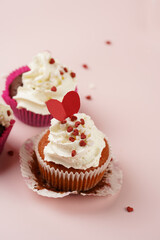 Three homemade white and chocolate muffins with white creamy frosting and heart-shared red and white sugar decoration on marble board on pink surface