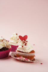 Three homemade white and chocolate muffins with white creamy frosting and heart-shared red and white sugar decoration on marble board on pink surface