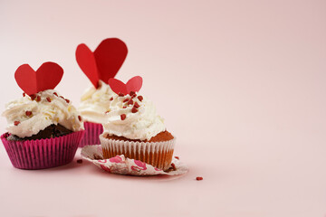 Three homemade white and chocolate muffins with white creamy frosting and heart-shared red and white sugar decoration on marble board on pink surface