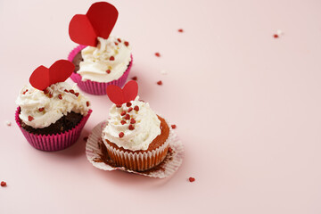 Three homemade white and chocolate muffins with white creamy frosting and heart-shared red and white sugar decoration on marble board on pink surface