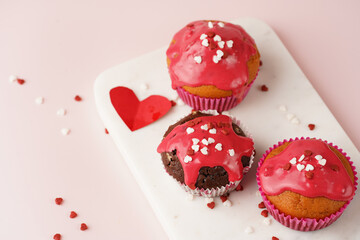 Three homemade white and chocolate muffins with red frosting and heart-shared red and white sugar decoration on marble board on pink surface