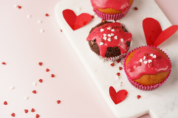 Three homemade white and chocolate muffins with red frosting and heart-shared red and white sugar decoration on marble board on pink surface