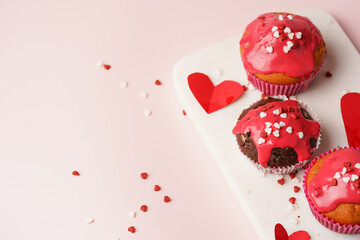 Three homemade white and chocolate muffins with red frosting and heart-shared red and white sugar decoration on marble board on pink surface