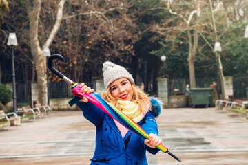 Young adult woman wearing blue jacket and yellow gloves, holding colorful umbrella in winter...