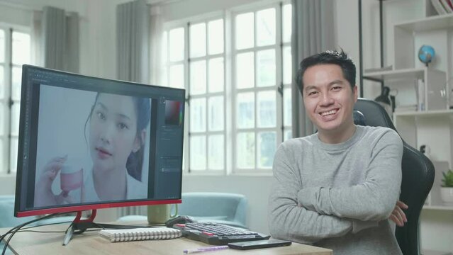 Asian Cameraman In Long Sleeved T-Shirt Crossing His Arms And Smiling To Camera While Editing Photo By Desktop Computer At Home.
