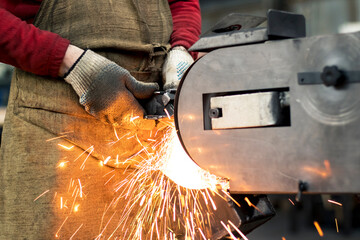 A skilled worker in gloves and an apron grinds a steel part on a belt grinder, a lot of sparks fly...