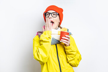 Young woman yawns with her mouth open wide while holding coffee in a paper cup on a light background.