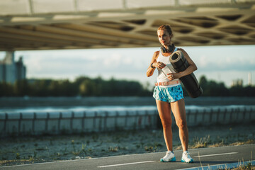 Woman With Rolled Up Exercise Mat And Water Bottle Resting After Training Near The River