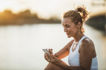 Woman Sitting Near The River And Using Smart Phone Before Training At The Sunset