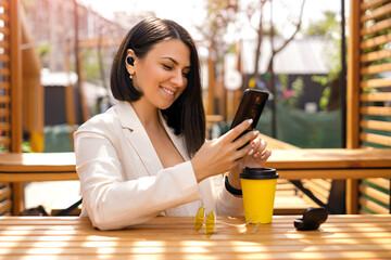 Brunette Caucasian business lady with small wireless black headphones in her ears looks at the phone with a smile, reading messages. Woman is relaxing in a cafe on the terrace enjoying music - obrazy, fototapety, plakaty