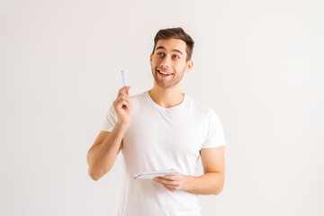 Studio portrait of enthusiastic young man writing in copybook, raising pen up eureka gesture on white isolated background. Happy male studying, preparing for exam, making notes in paper notebook.