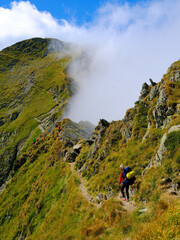 Fototapeta premium Young woman with a backpack descends the rocky terrain with a blurred view of the mighty green mountains and the blue sky with clouds. Fagaras Mountains, Carpathians, Romania
