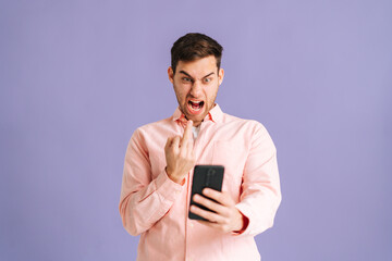 Portrait of angry young man showing middle finger to smartphone screen on pink isolated background. Studio shot of handsome male aggressively communicating on video call using mobile phone.