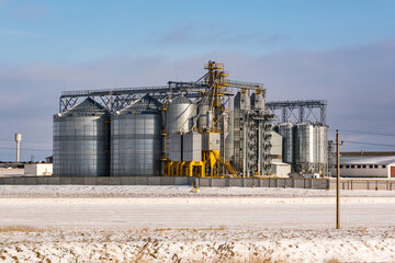 Obraz premium agro silos granary elevator in winter day in snowy field. Silos on agro-processing manufacturing plant for processing drying cleaning and storage of agricultural products, flour, cereals and grain.