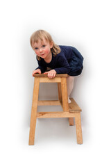 A two-year-old girl in a dark blue dress climbs onto a two-step chair. He stands on the first step and looks into the camera. Blonde with blue eyes. On a white background.