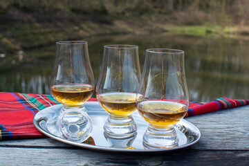 Tasting glasses of scotch whisky strong drink and red Scottisch tartan on old outdoor table with forest lake on background