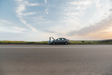 A young girl stands near a broken-down car in the middle of the highway during sunset and tries to call for help on the phone. Waiting for help. Car service. Car breakdown on road.