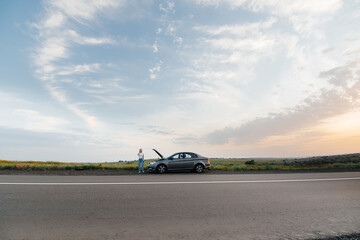 A young girl stands near a broken-down car in the middle of the highway during sunset and tries to call for help on the phone. Waiting for help. Car service. Car breakdown on road.