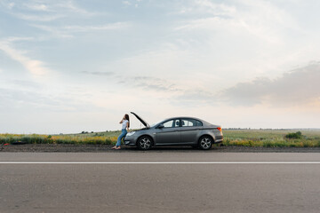 Obraz na płótnie Canvas A young girl stands near a broken-down car in the middle of the highway during sunset and tries to call for help on the phone. Waiting for help. Car service. Car breakdown on road.