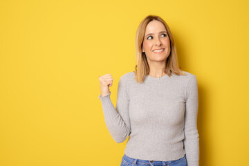 Smiling woman in grey sweater showing thumb up standing isolated over yellow background.