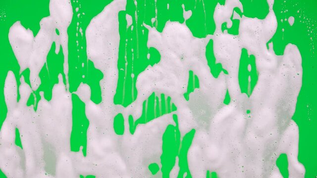 White soap suds flowing down window glass on green screen chroma key background. Cleaning windows with cleaning disinfectant. Hygiene, disinfection, housework, household, cleanup, washing. Close up.