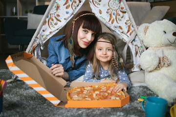 Beautiful mother and her loving daughter eating fresh tasty pizza while playing inside teepee tent...