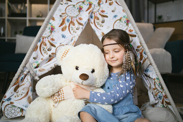 Lovely little child sitting near teepee tent in embrace with her soft white teddy bear. Happy...