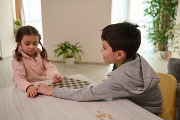 Two adorable European elementary aged kids, boy and girl, brother and sister having great time playing checkers board game together at home interior