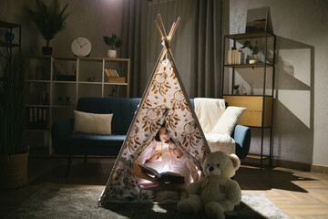 Caucasian mother and her pretty daughter sitting in wigwam and reading book during evening time using flashlight. Cozy home and family concept. © sofiko14