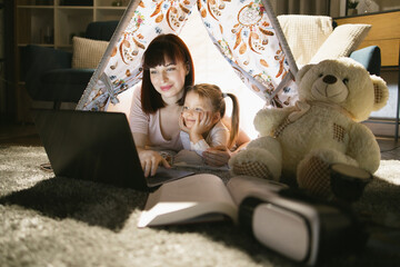 Beautiful caucasian woman and pretty female child lying together in toy wigwam and using modern laptop during evening time. Technology at home. © sofiko14