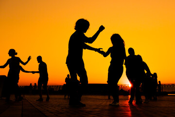 Woman and man couple silhouettes dancing against warm sunset orange sky on quay at evening. Group dance, romantic, love, summer outdoor activity and vacation concept
