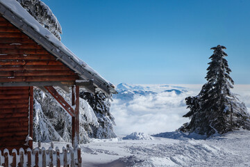 Beautiful Winter Landscape With Log Cabin,  Mountain Peaks In The Distance, Conifer Tree And Blue Sky.