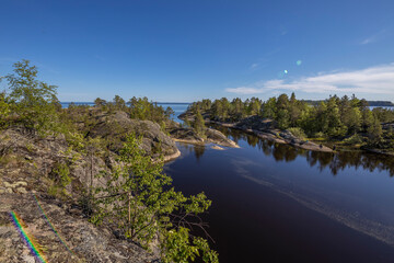 Landscape with a forest on stones over the lake. Sunny day at the lake. Reflection of the sky in the water. Pines on stones. The nature of the north.