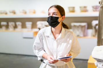 Portrait of young female visitor in protective face mask with paper guide at paintings exposition