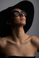 Dark studio fashion portrait of elegant young and sexy woman in black wide hat, black classic evening dress and sunglasses. Stylish elegant woman look aside of camera in grey background