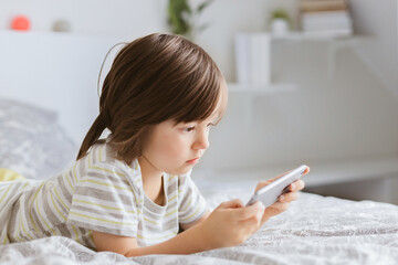 Charming boy lies on the bed and uses a mobile phone. The child plays online games. Children and...