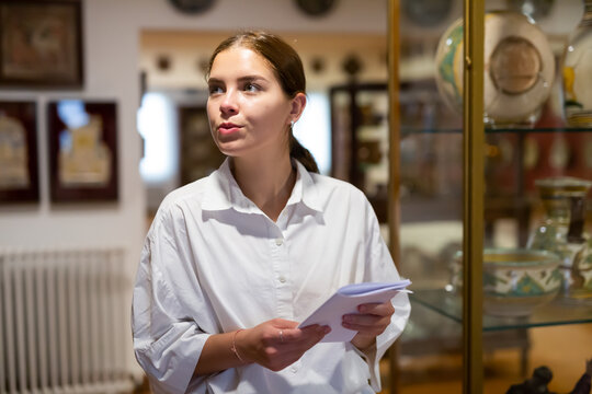 Young attractive female with information brochure looking at the exposition of antique ceramic at historic museum