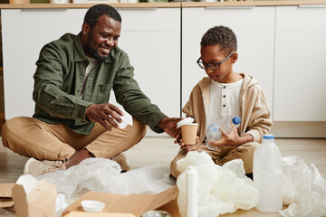 Full length portrait of happy African-American father and son sorting plastic at home for waste...